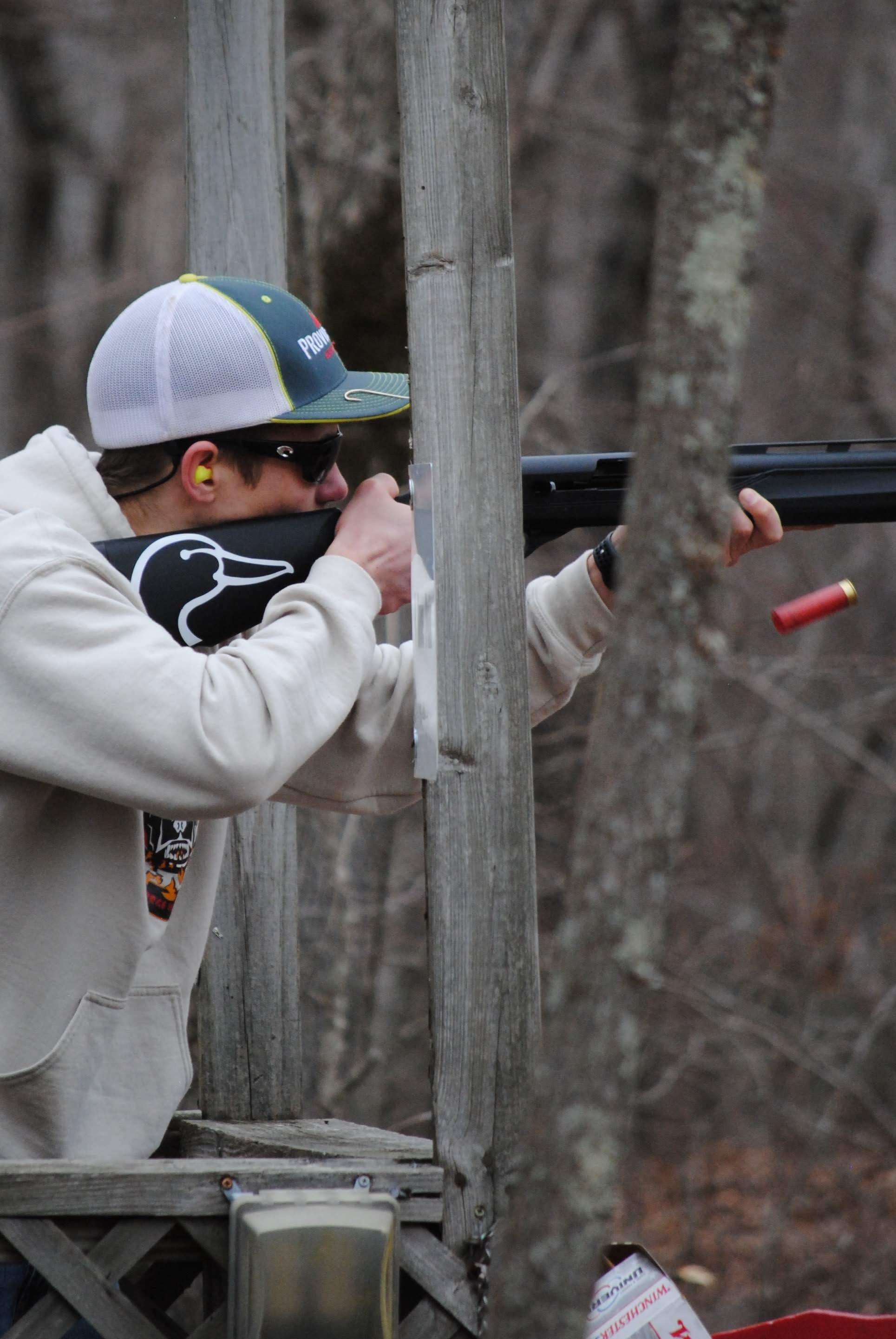 Youth shooting a shotgun, wearing eye and ear protection.  Shotgun shell caught in the air after being ejected from the gun.